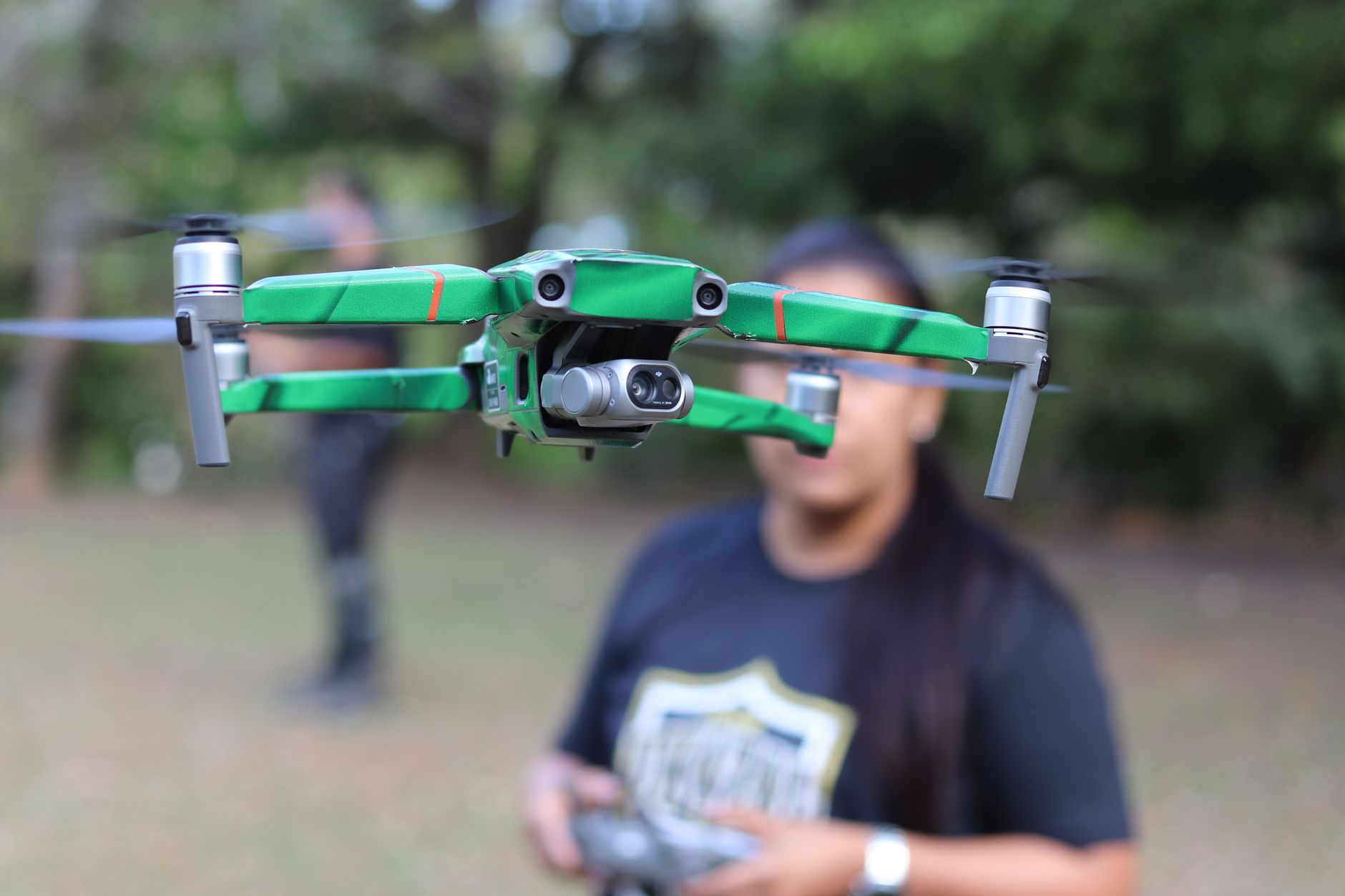 Drone pilot holding a controller while a small drone hovers above a grassy field
